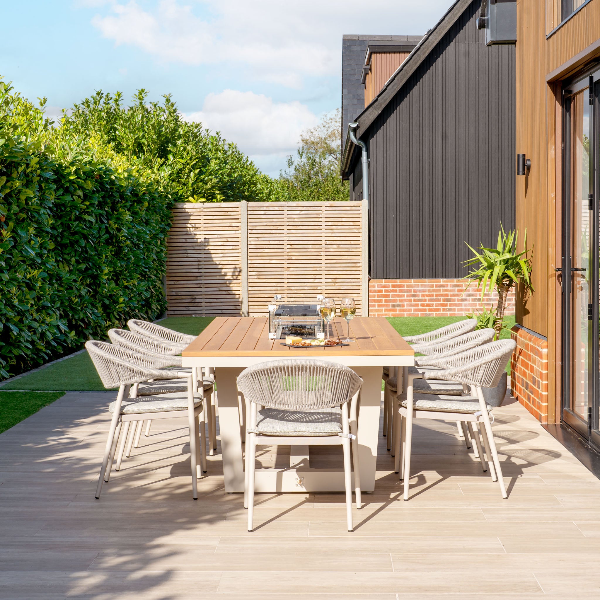 A Cloverly 8 Seat Rectangular Teak Firepit Dining Set in Latte, complemented by a grill and surrounded by lush greenery, creates the perfect outdoor setting for relaxation.