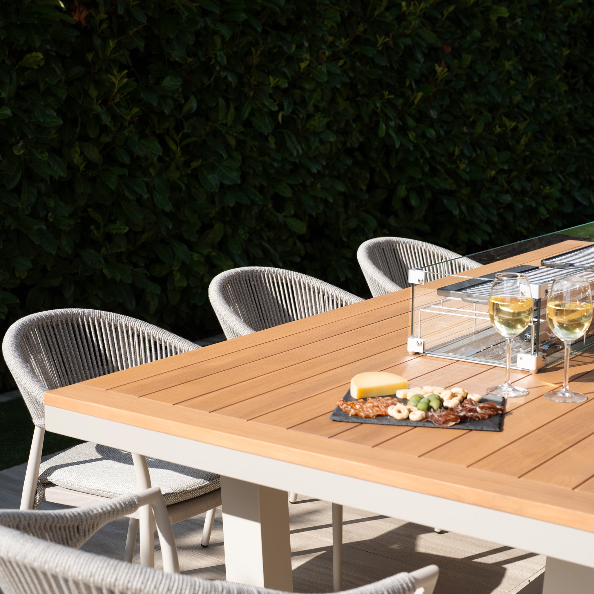 An outdoor dining setup featuring the Cloverly 8 Seat Rectangular Teak Firepit Dining Set in Latte, complete with a wooden table, cheese platter, and wine glasses, surrounded by lush green foliage. UV-resistant chairs enhance the practicality of the scene.