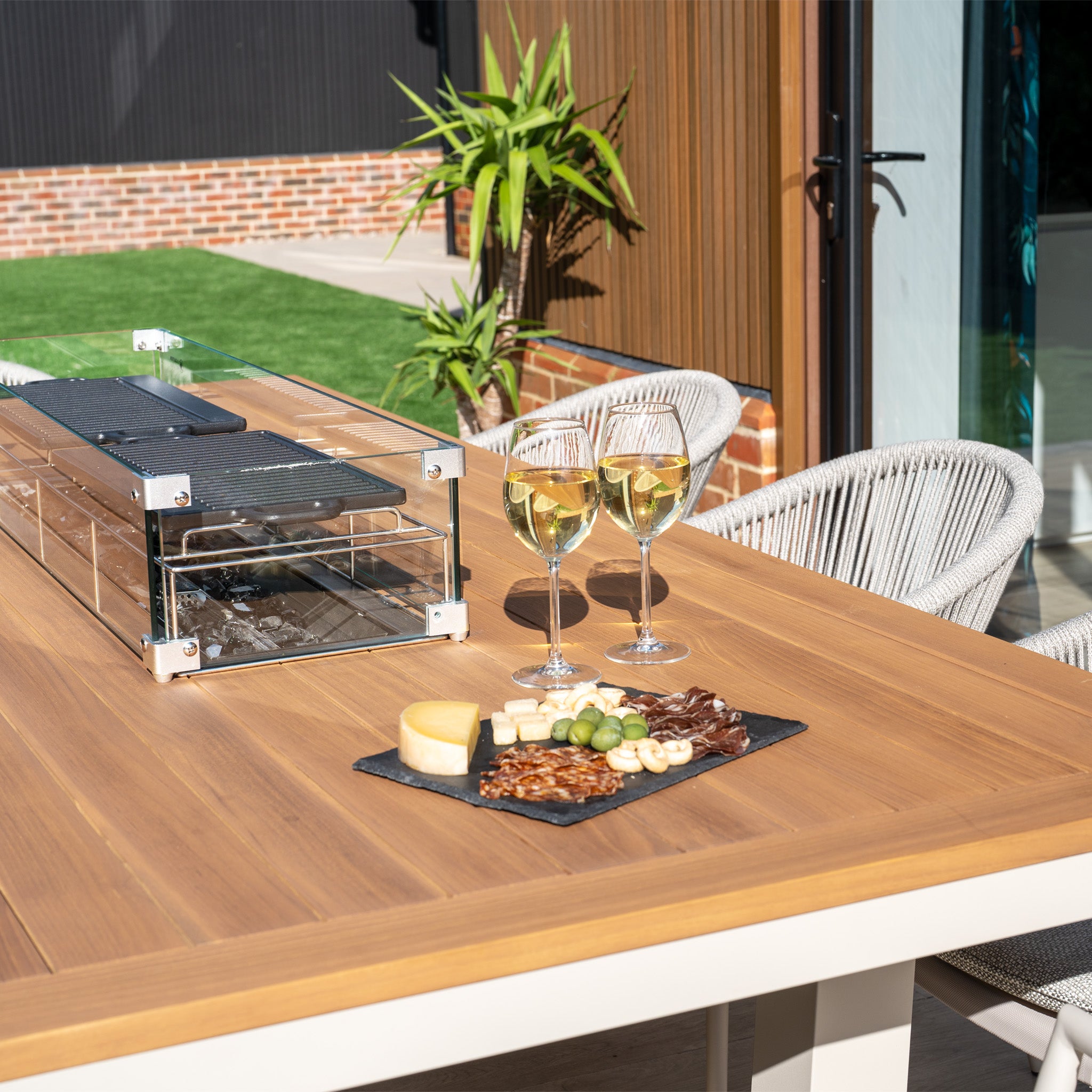 Cheese and wine elegantly rest on an outdoor table surrounded by UV-resistant chairs, complemented by a grill in the sunny grassy yard. The scene is perfect for enjoying cozy evenings as the Cloverly 8 Seat Rectangular Teak Firepit Dining Set in Latte adds warmth to every gathering.