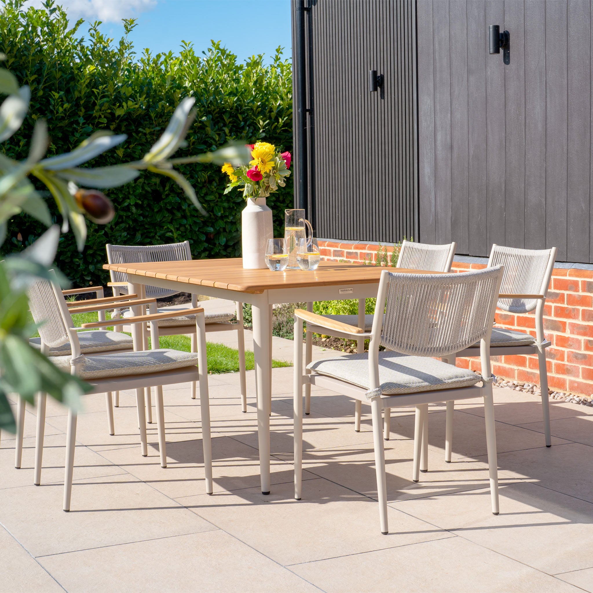 The Tellaro 6 Seat Rectangular Dining Set with Teak Table in Latte embodies outdoor living, featuring a teak table and six chairs, enhanced by a floral vase with a leafy backdrop and brick wall.