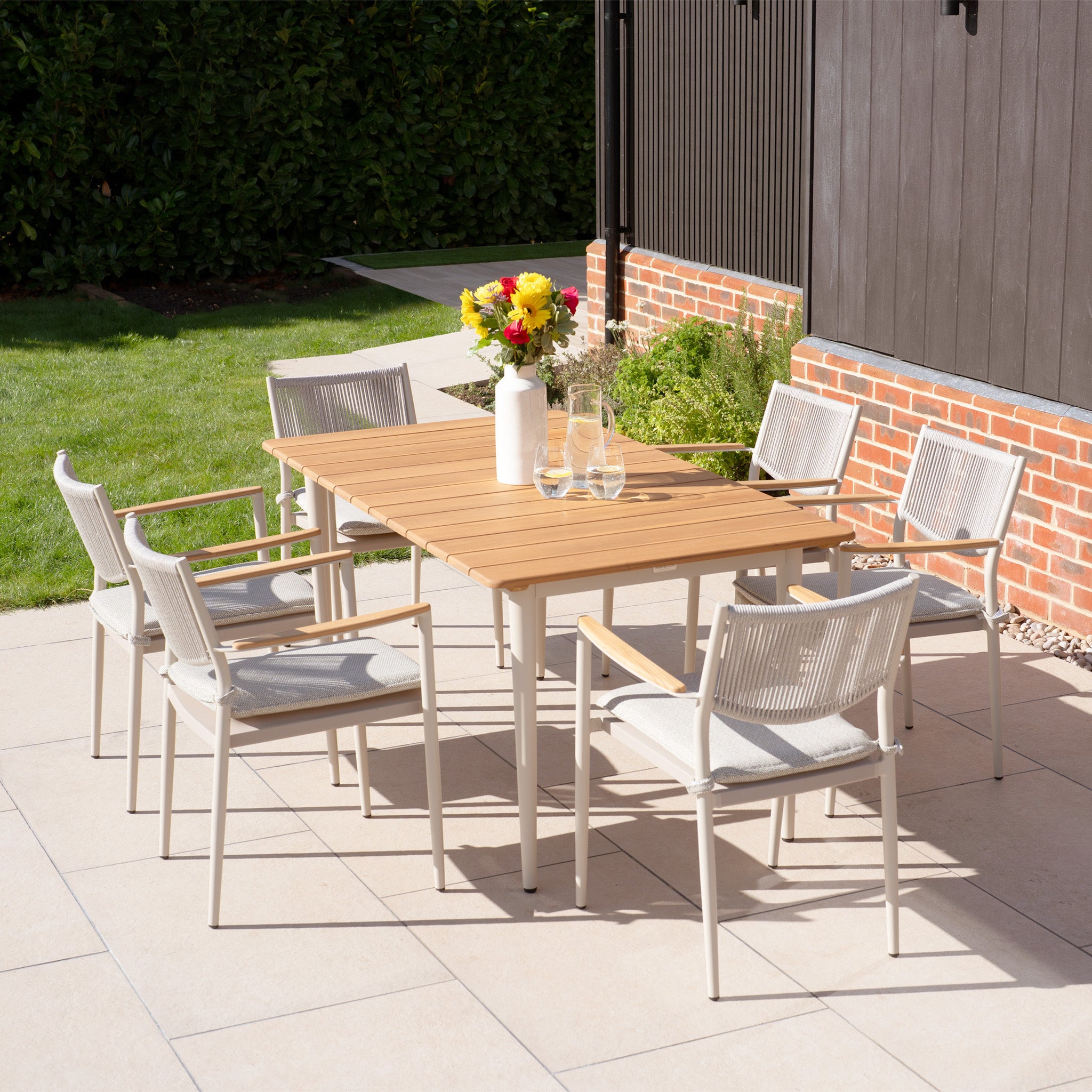 The Tellaro 6 Seat Rectangular Dining Set with a teak table in Latte is ideal for outdoor living, featuring six chairs and a vase of flowers on a patio near the green lawn and brick wall.