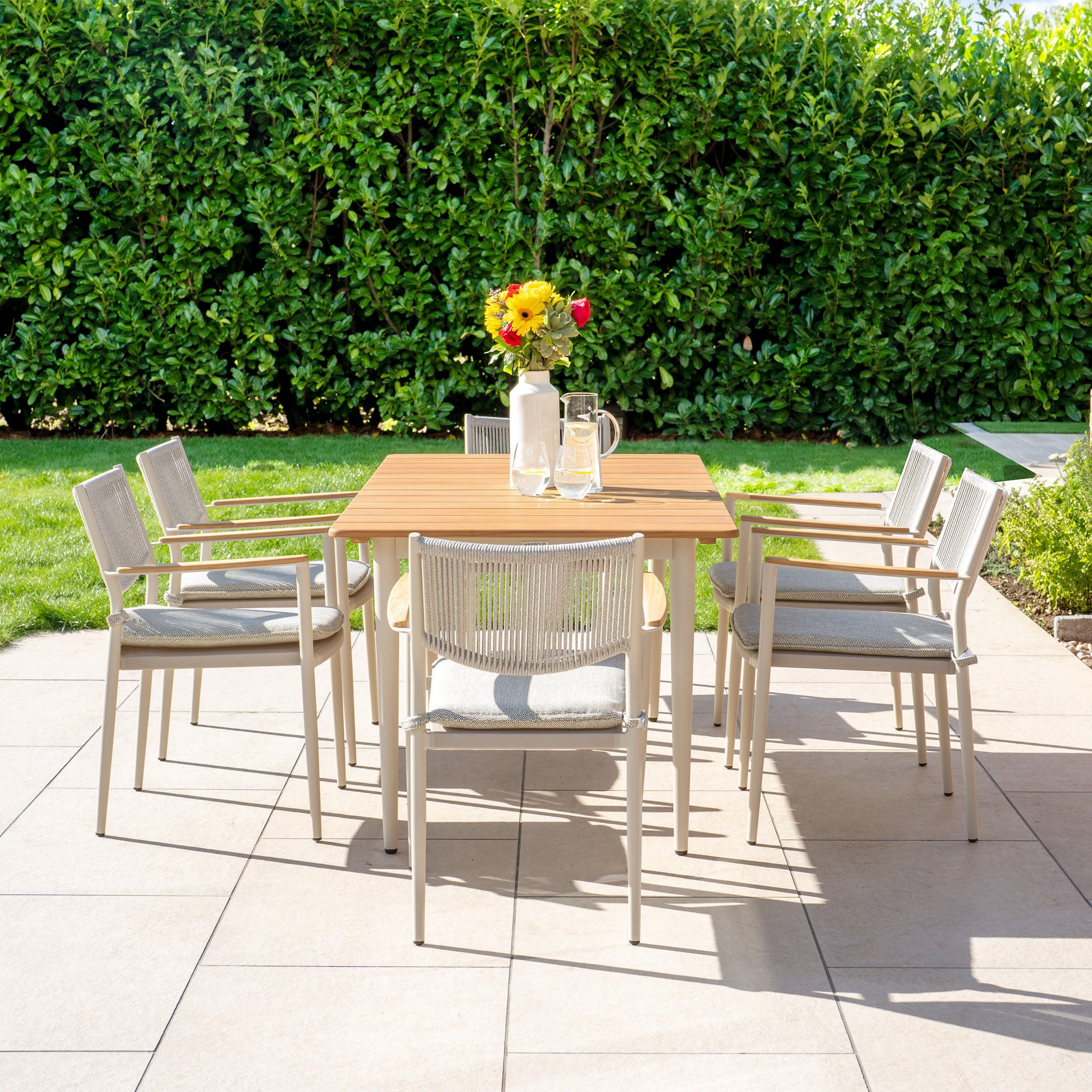 The Tellaro 6 Seat Rectangular Dining Set with a Teak Table in Latte is ideal for outdoor living, offering a stylish setup with six chairs and natural elegance amidst lush greenery.