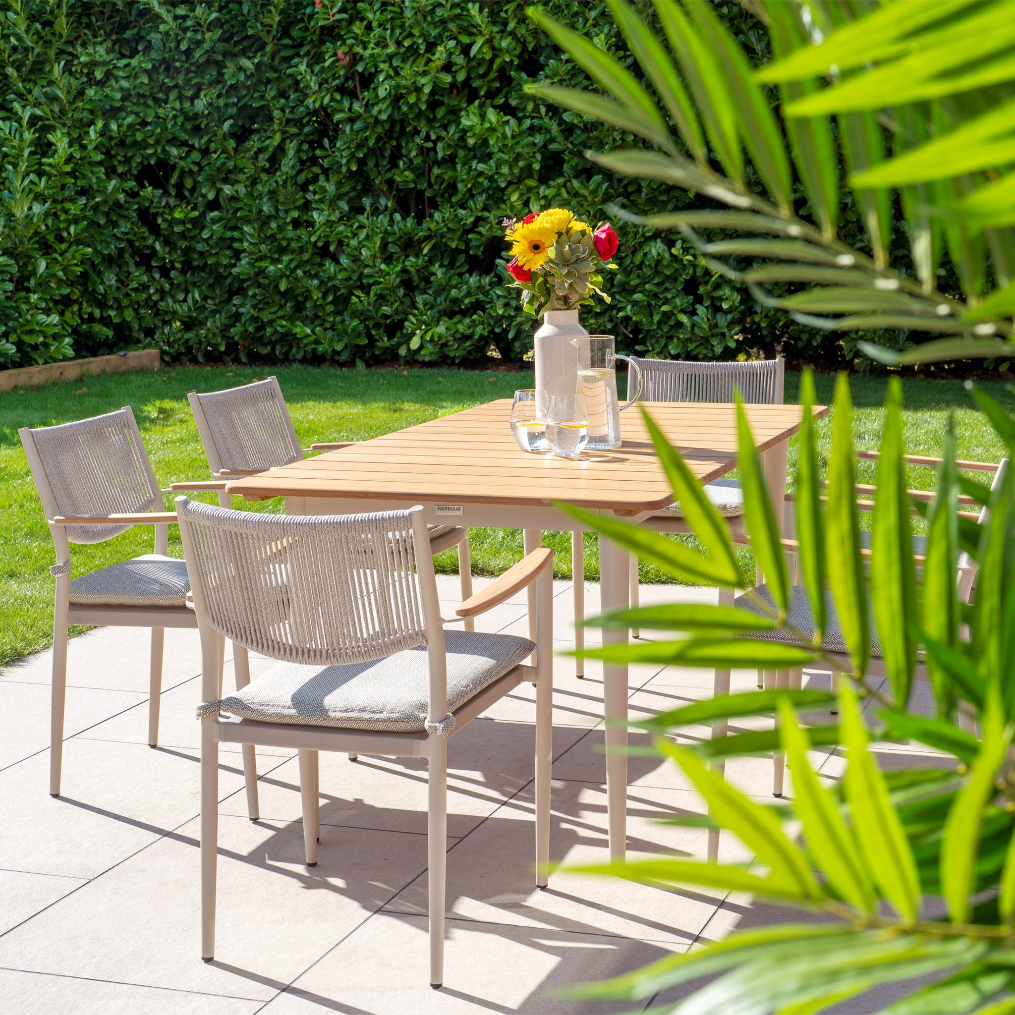 Experience outdoor living on a sunny patio featuring the Tellaro 6 Seat Rectangular Dining Set with a Teak Table in Latte. Surrounded by lush green plants, it's ideal for relaxation and al fresco dining, complete with a charming vase of flowers.