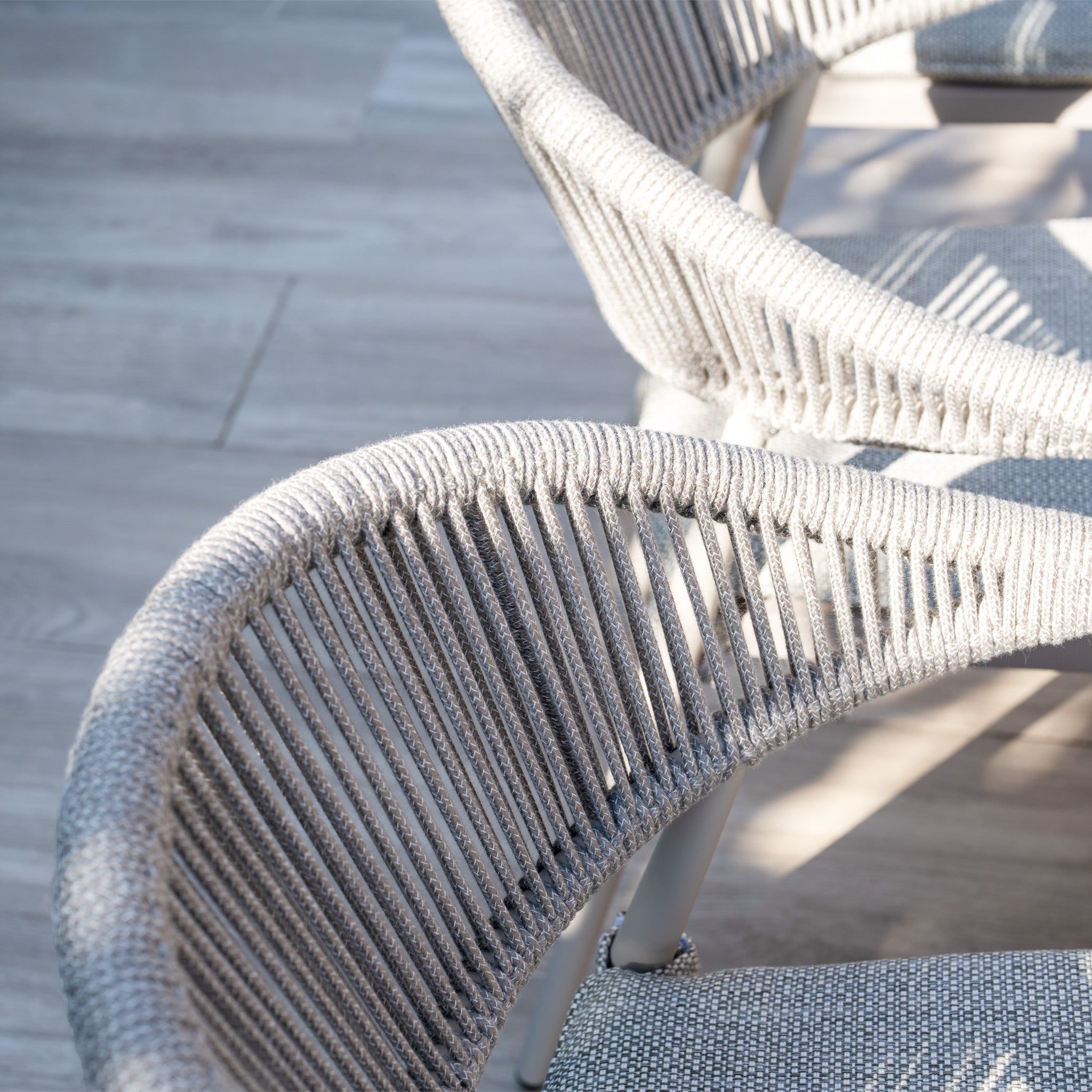 Close-up of woven gray chairs with curved backs on a wooden floor, partially lit by sunlight, as part of the elegant Cloverly 8 Seat Rectangular Teak Firepit Dining Set in Latte.