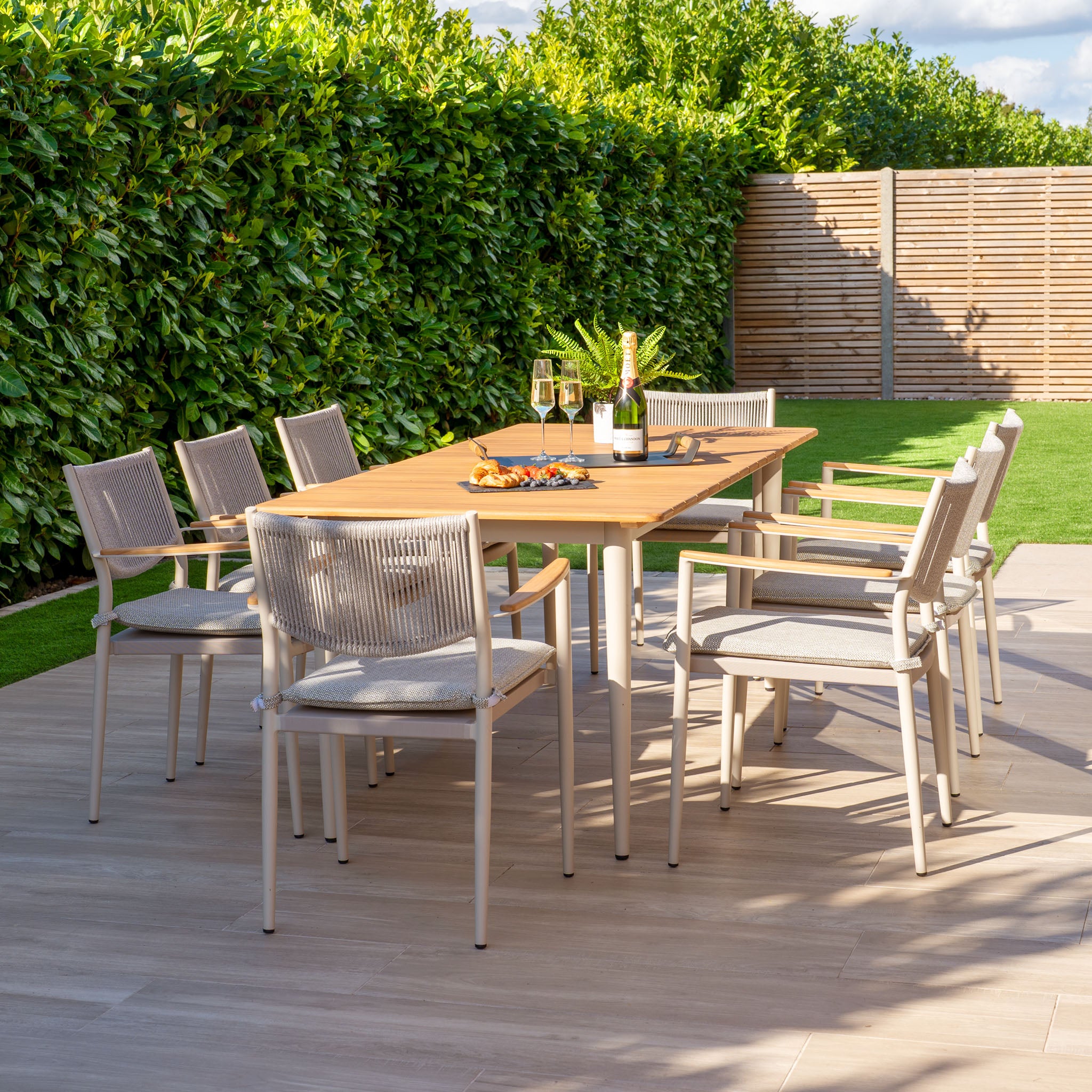 The Tellaro 8 Seat Rectangular Dining Set features a weather-resistant teak table in Latte, surrounded by lush greenery and enclosed by a wooden fence.