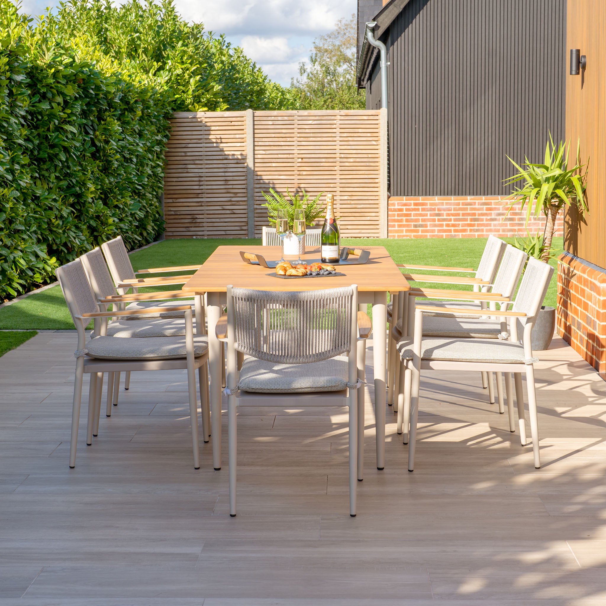 Outdoor dining is a delight with the Tellaro 8 Seat Rectangular Dining Set featuring a teak table in Latte, weather-resistant chairs, lush plants, and a wooden privacy fence in a charming garden setting.