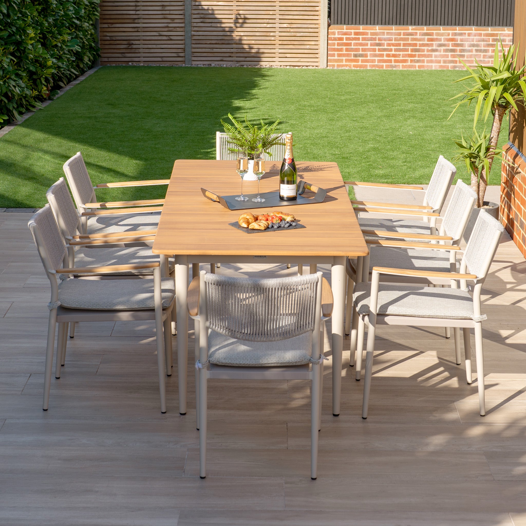 The Tellaro 8 Seat Rectangular Dining Set in Latte, featuring a teak table, is displayed with wine and snacks on a patio. Surrounded by lush greenery, its weather-resistant design ensures style and durability for any gathering.