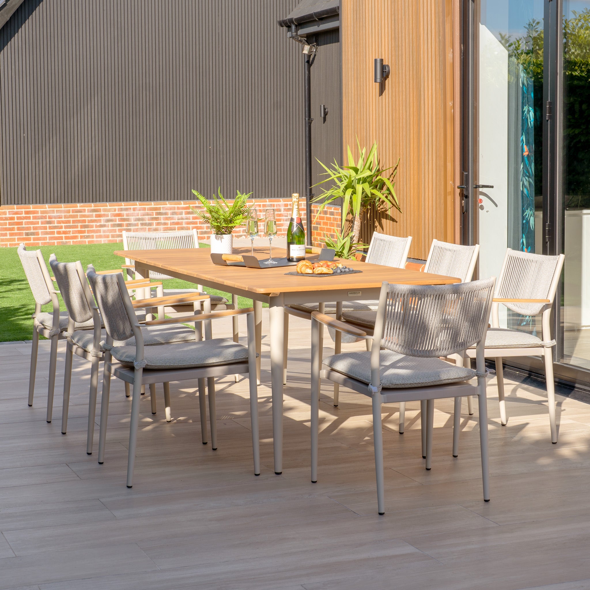 The patio features the Tellaro 8 Seat Rectangular Dining Set with a weather-resistant teak table in Latte, surrounded by lush plants and a bottle.