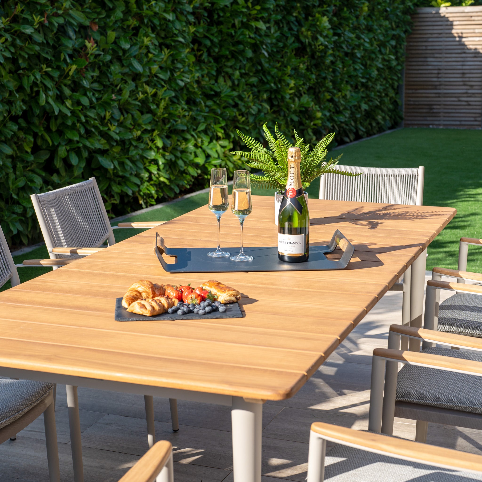 Enhance outdoor dining with the weather-resistant Tellaro 8 Seat Rectangular Dining Set with a teak table in Latte, beautifully styled with champagne, croissants, berries, and leafy decor in a garden setting.