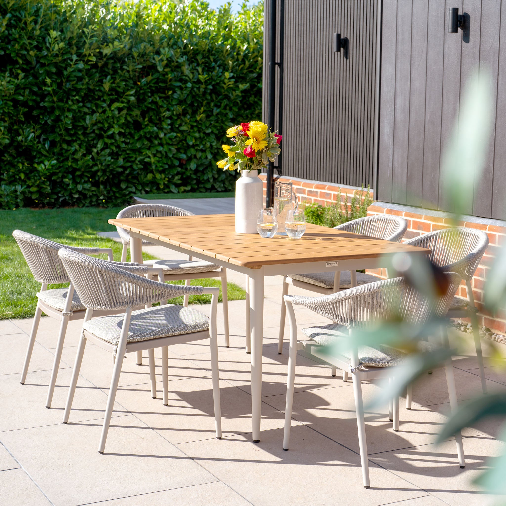 The Cloverly 6 Seat Rectangular Dining Set in Latte includes a wooden table with a teak top, encircled by six chairs that enhance the elegance of the patio. Its aluminum frame beautifully complements the natural setting, while a vase of flowers adds charm against the lush greenery backdrop.