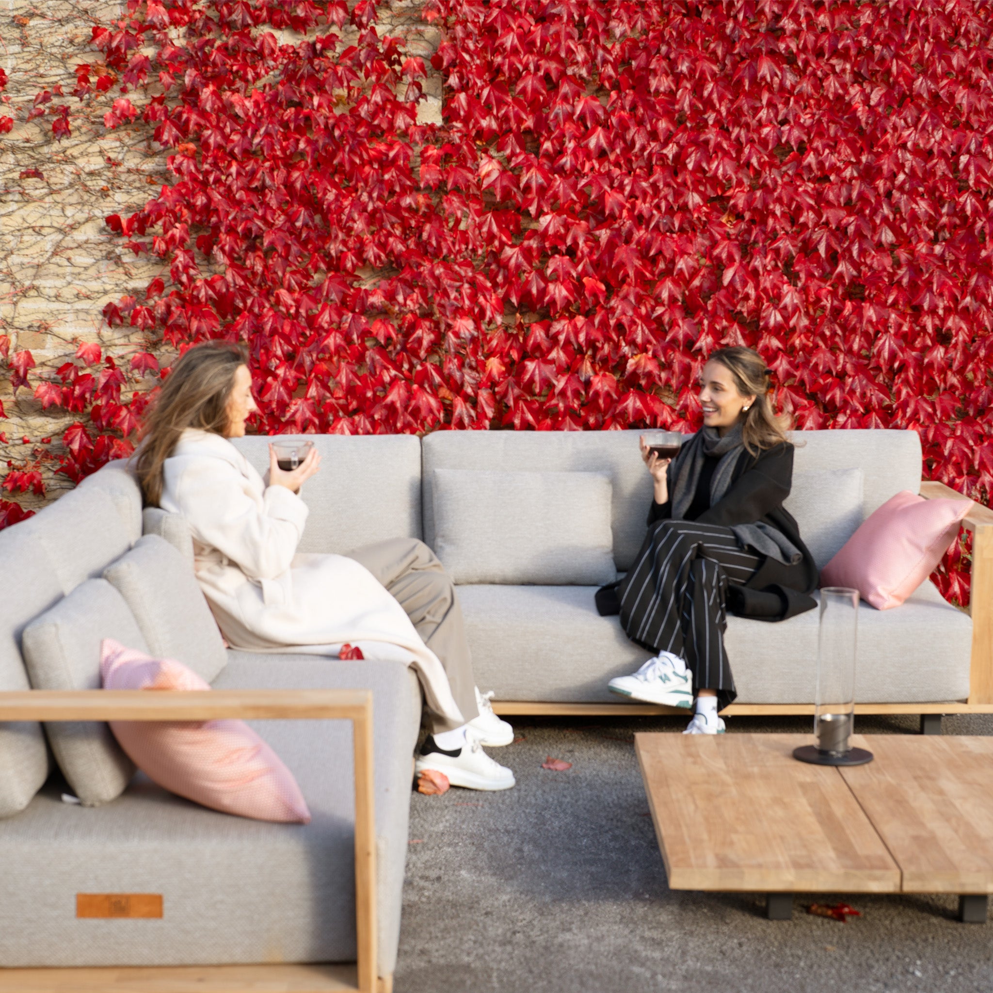 Two people sit on outdoor sofas with red ivy on the wall, holding drinks, smiling, and conversing comfortably. They enjoy their serene setting surrounding the Eternity Right Hand Teak Corner Group in Light Grey, crafted from elegant teak wood.