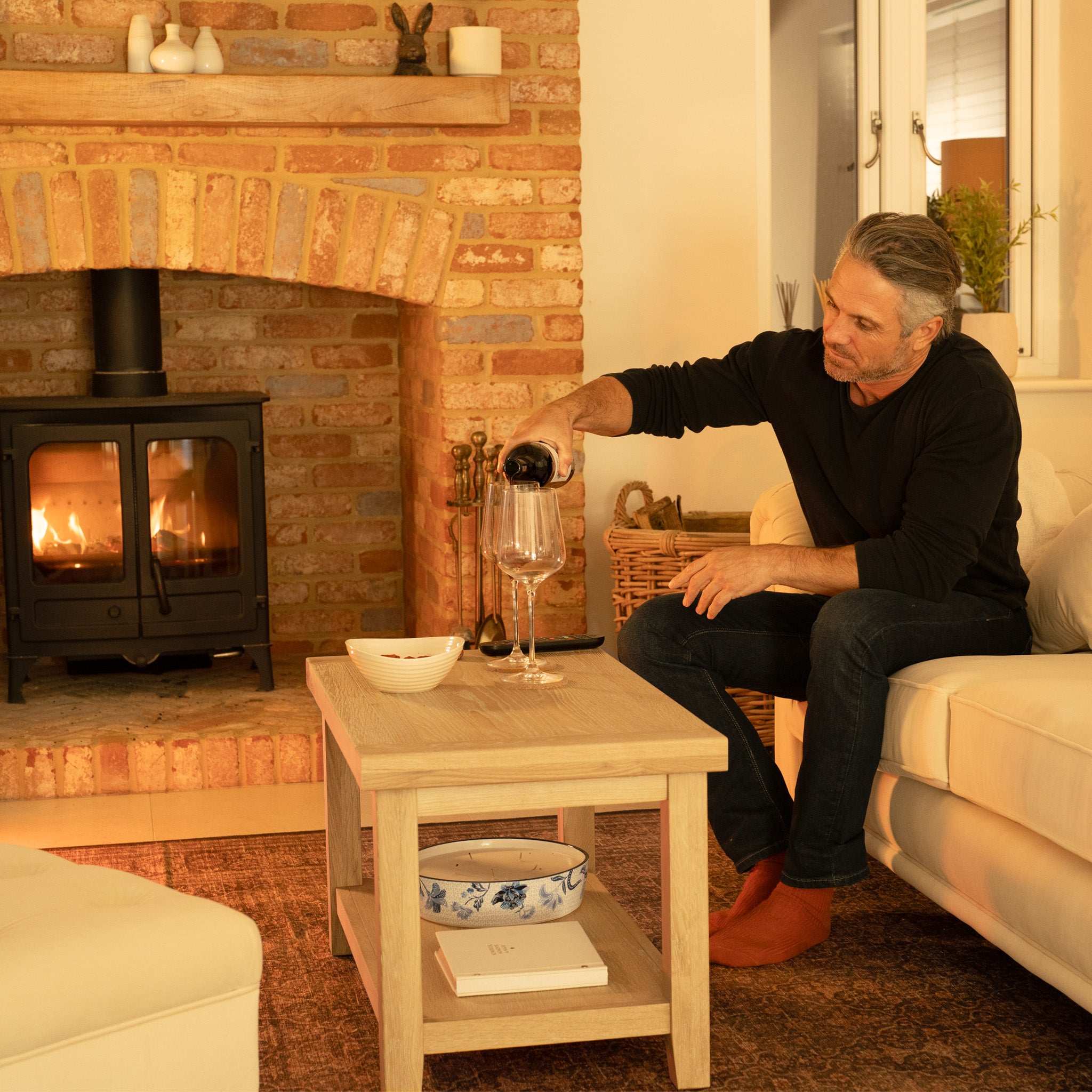 A man pours wine in a cozy living room with a fireplace and brick decor, beside the Burford Large Coffee Table with Shelf in Natural Oak.