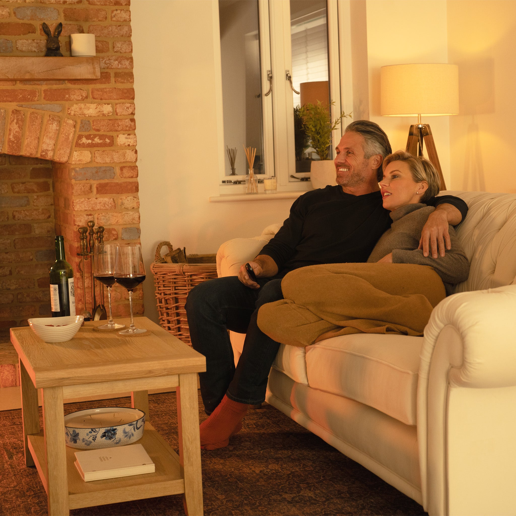 A couple relaxes on a sofa by the fireplace, wrapped in a blanket with wine glasses on the Burford Large Coffee Table with Shelf in Natural Oak, its elegant finish adding charm to their cozy evening.