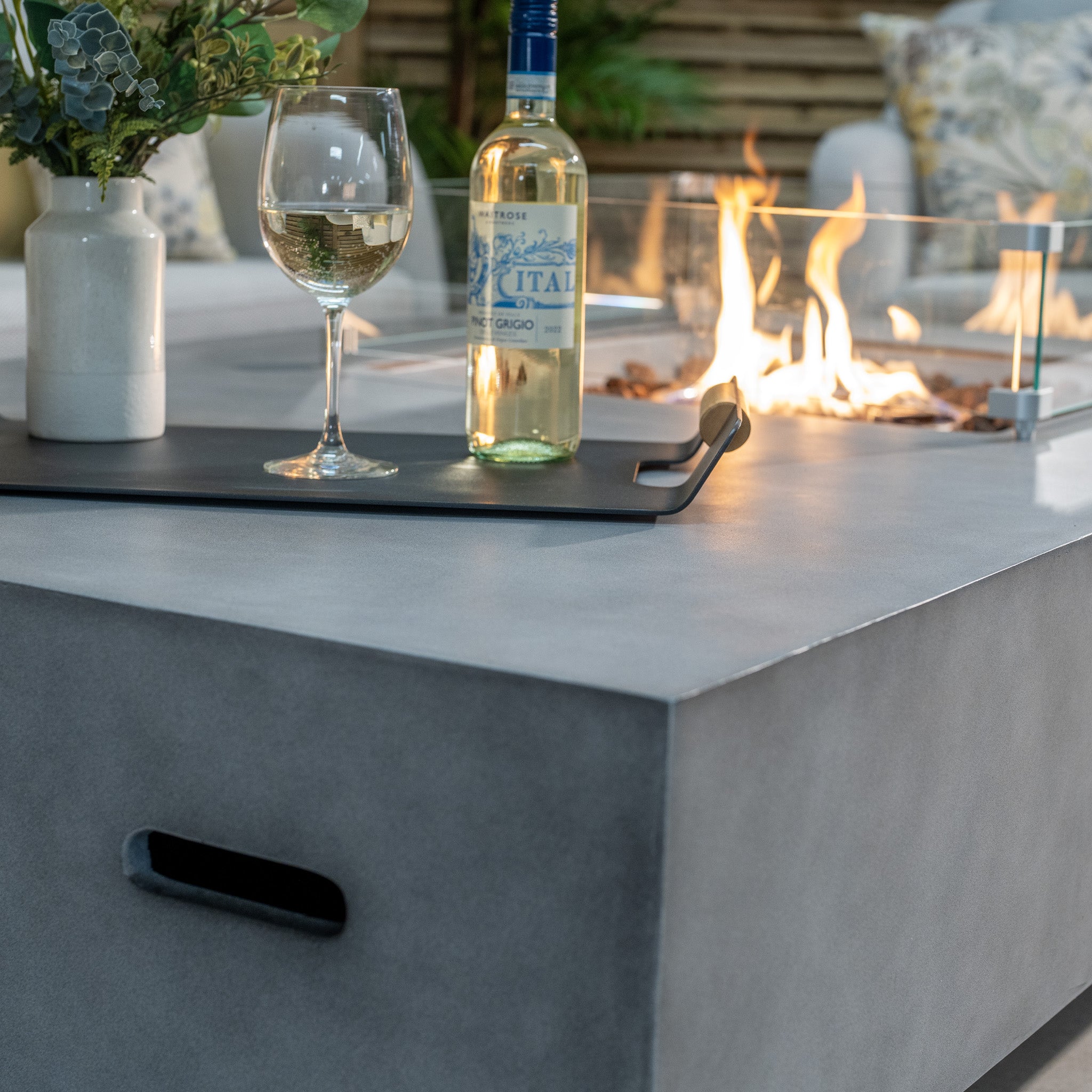 Ember Rectangular Firepit Coffee Table in Stone Grey with a glass of wine, wine bottle, flowers, and a lit fire glass barrier. Crafted from 304 stainless steel and boasting a 14kW heat output, it's the perfect centerpiece for any outdoor gathering.