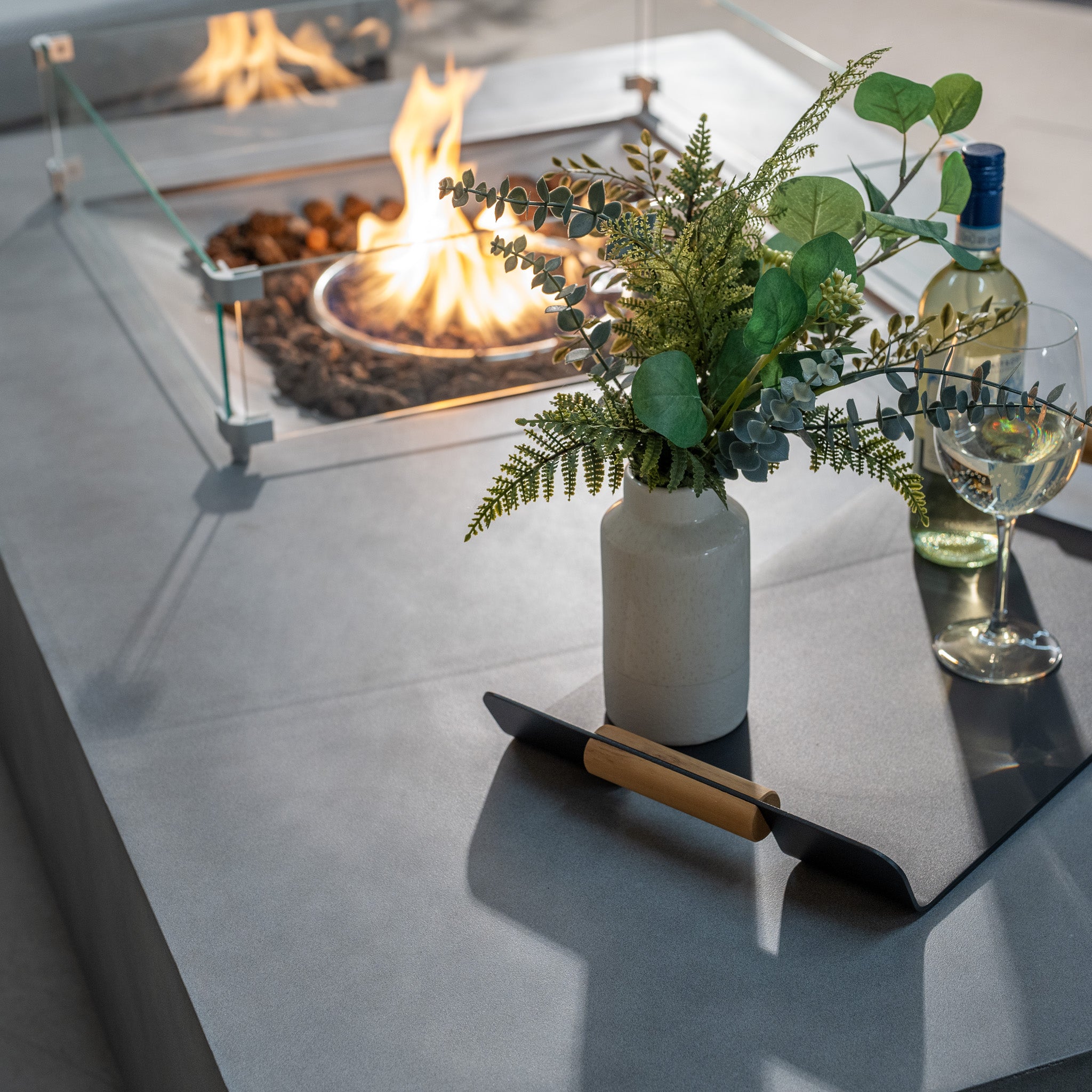 The Ember Rectangular Firepit Coffee Table in Stone Grey, crafted from 304 stainless steel, showcases a small fire pit with an impressive 14kW heat output. A black tray holds a vase of greenery, a wine bottle, and a glass on this elegant piece.