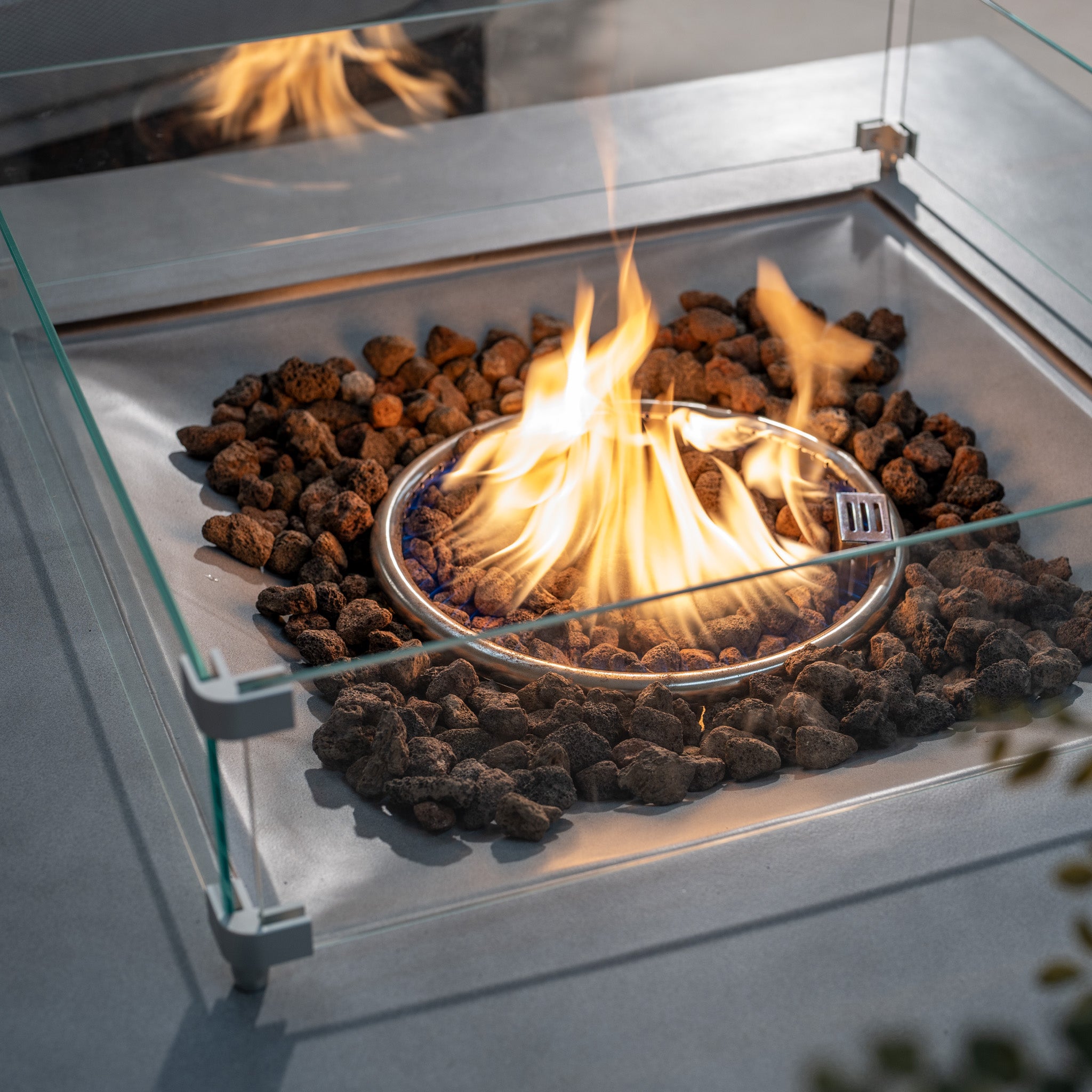 The Ember Rectangular Firepit Coffee Table in Stone Grey features glass sides and burns a small fire over a bed of lava rocks. Made from durable 304 stainless steel, it offers a cozy ambiance with an impressive 14kW heat output.