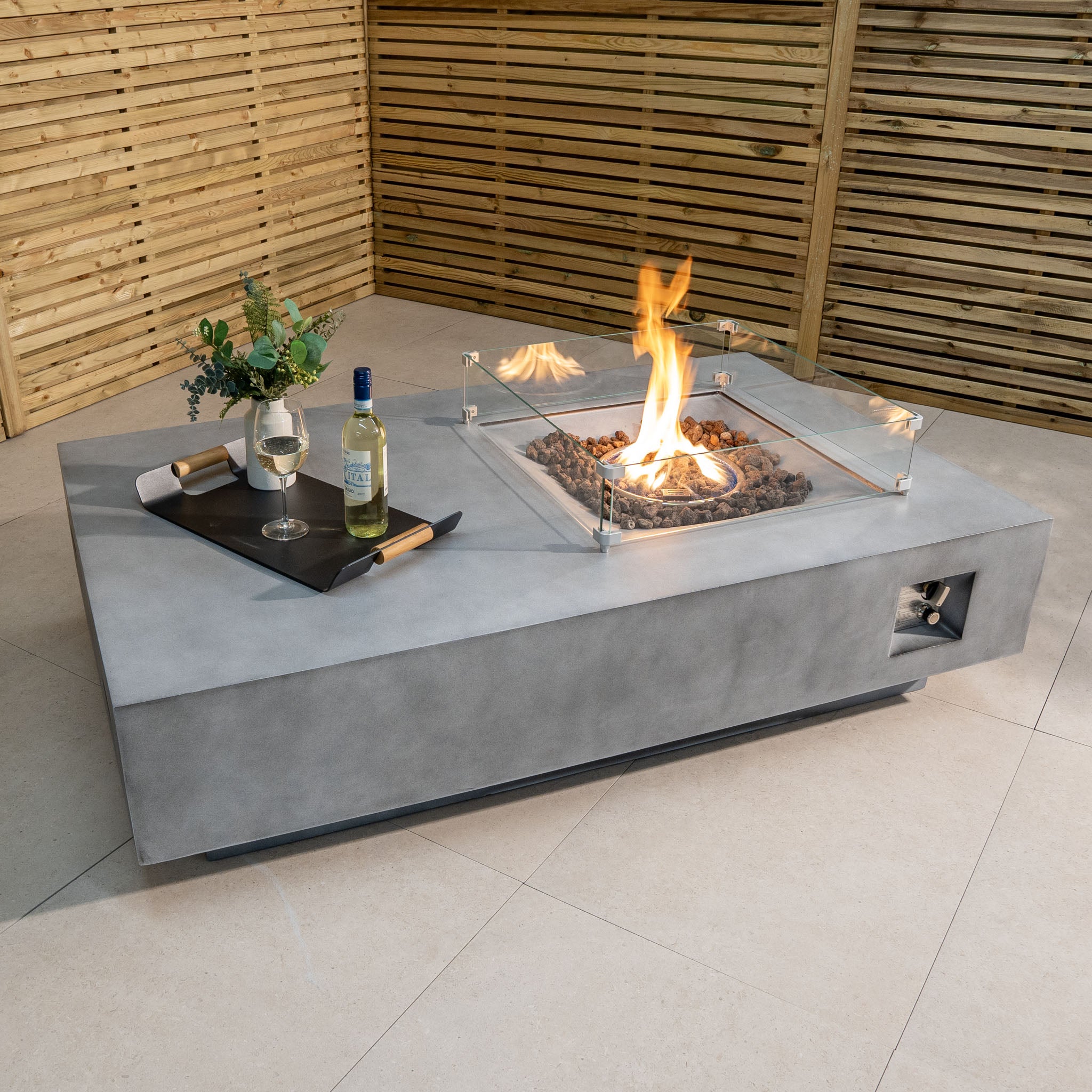 Experience cozy evenings with the Ember Rectangular Firepit Coffee Table in Stone Grey, featuring a modern concrete design, glass walls, and a 14kW heat output. Enjoy a lit fire with a wine bottle and glasses on a tray in your wooden-fenced patio for the perfect ambiance.