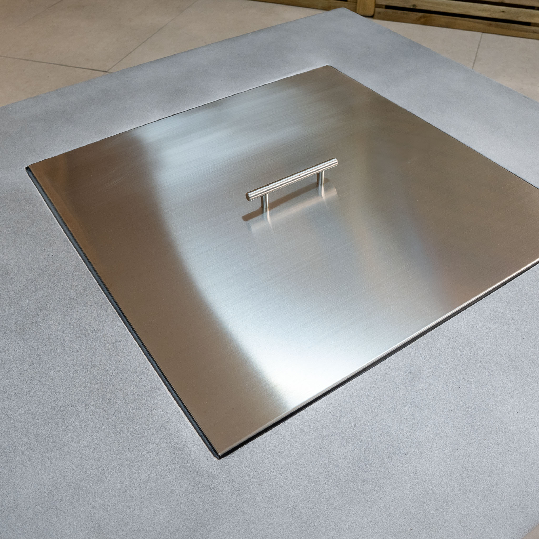 A square 304 stainless steel cover with a handle rests on a smooth, light gray surface, perfectly complementing the Ember Rectangular Firepit Coffee Table in Stone Grey with its sleek design and 14kW heat output.