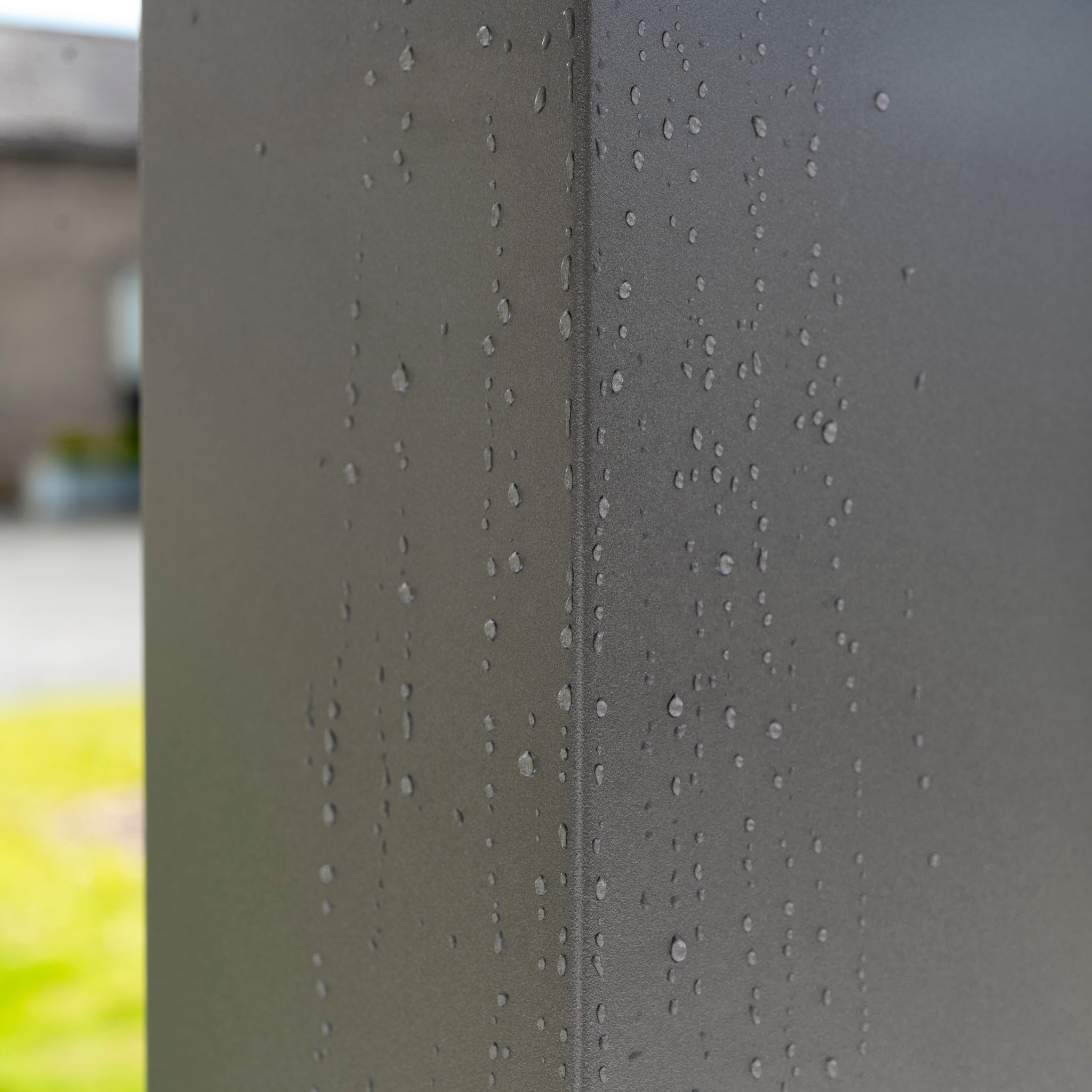 Close-up of water droplets on the sleek surface of a PergoSTET Deluxe Frame Only 3m x 9m Rectangular Pergola with Motorised LED Louvres in Grey, set outdoors against a backdrop of blurred greenery and a building.