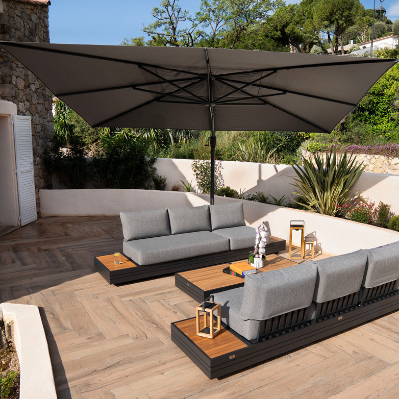 Outdoor patio with grey sectional sofas, a large Hacienda 3m x 4m Cantilever Parasol With Granite Base & Cover in Charcoal featuring a 360-degree rotation and UV-resistant canopy, wooden lanterns, and lush greenery in the background.