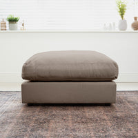 Cocoa Hampshire Modular Footstool with a plush cushion, placed on a patterned rug in a bright room with plants and décor on the window ledge; crafted from sustainably sourced hardwood for contemporary living.