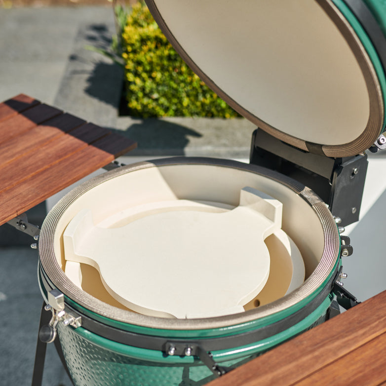 A ceramic Kamado grill with side shelves open, showcasing its green exterior and white interior components, including the Kamado Heat Deflector for the 22" Grill, stands proudly on a patio near bushes.