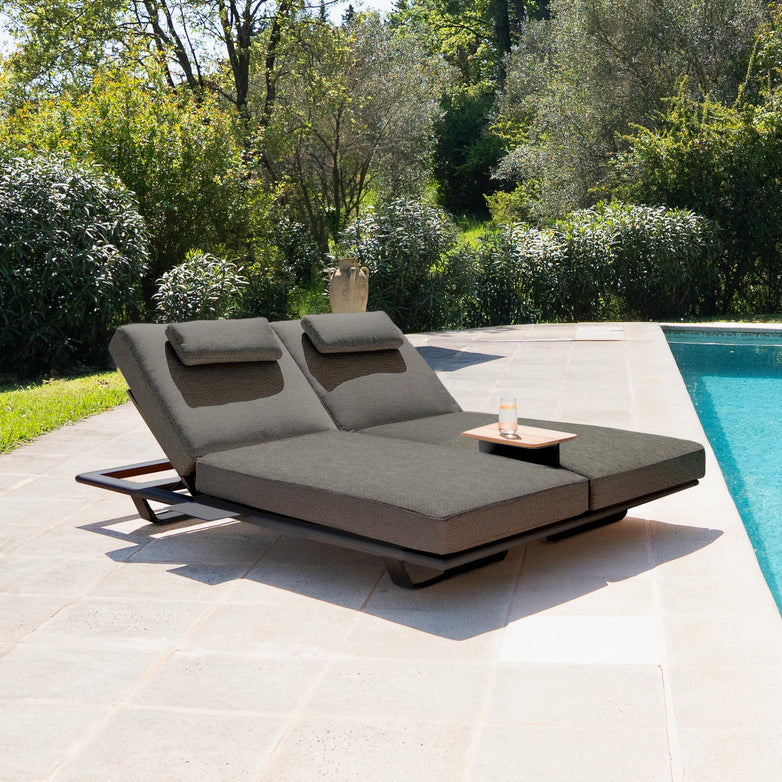 Two charcoal Hatia Double Sun Loungers with Side Tables, made with high-quality Olefin material and paired with plush pillows, sit beside a pool. Surrounded by greenery and trees in a sunny outdoor setting, these loungers offer both comfort and elegance for your relaxation needs.