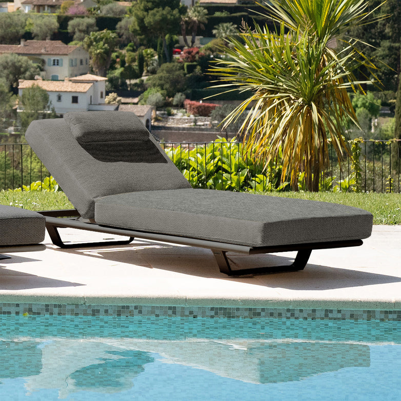 The Hatia Single Sun Lounger with Side Table in Charcoal, featuring an olefin material surface and a powder-coated aluminum frame, sits beside a clear blue pool surrounded by lush greenery, with houses in the background.