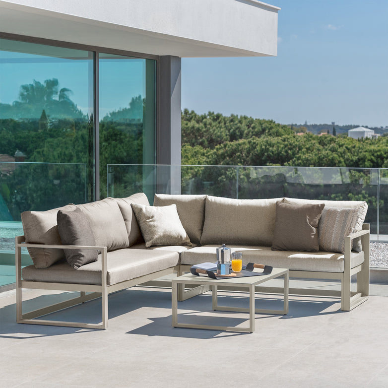 Modern outdoor patio featuring a Havana Corner Group Set with Reclining Feature and Coffee Table in Latte, showcasing a beige sectional sofa with pillows, a glass-top table, juice, and lush greenery in the background. This sleek outdoor living space provides the ultimate comfort and style.