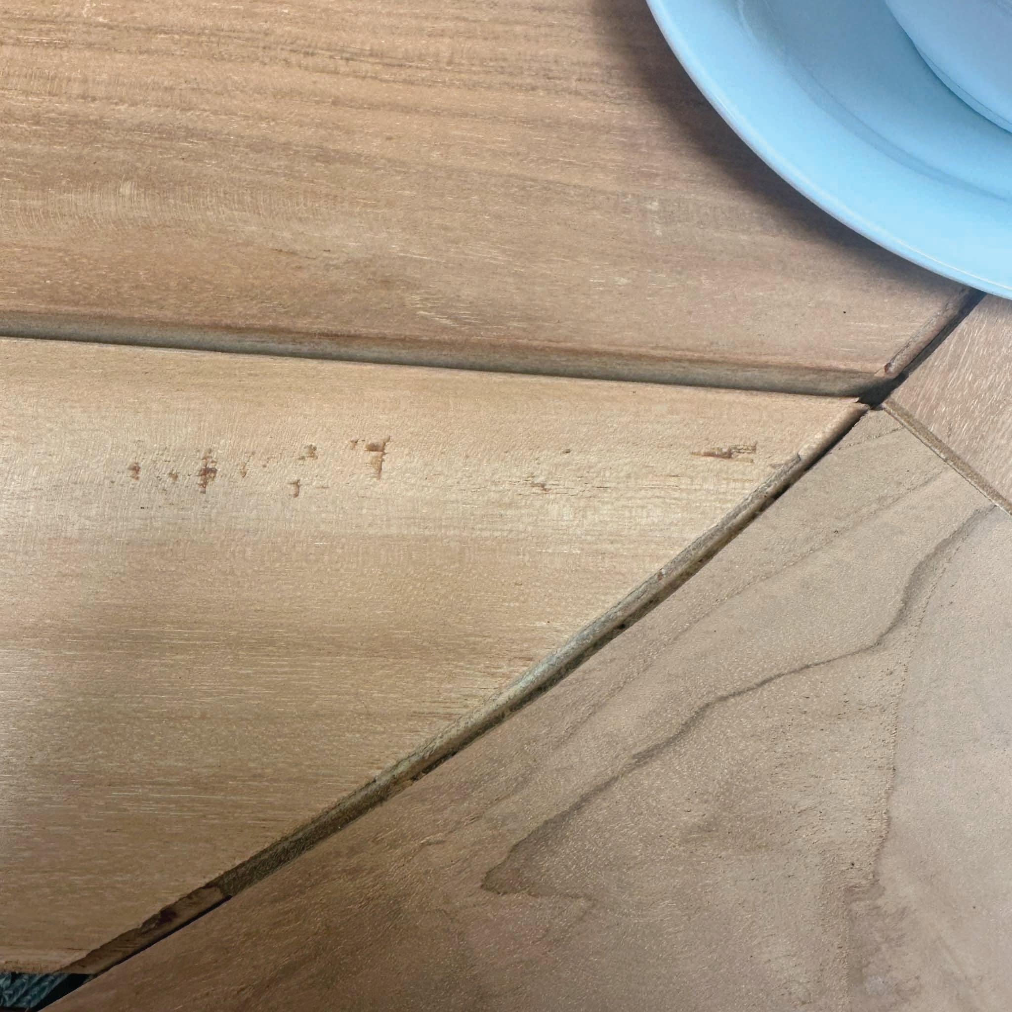 A close-up of a wooden table with light scratches and a partially visible white plate from the Bernini 6 Seat Round Dining with Teak Table in Light Grey (Ex Display) range in the top right corner.