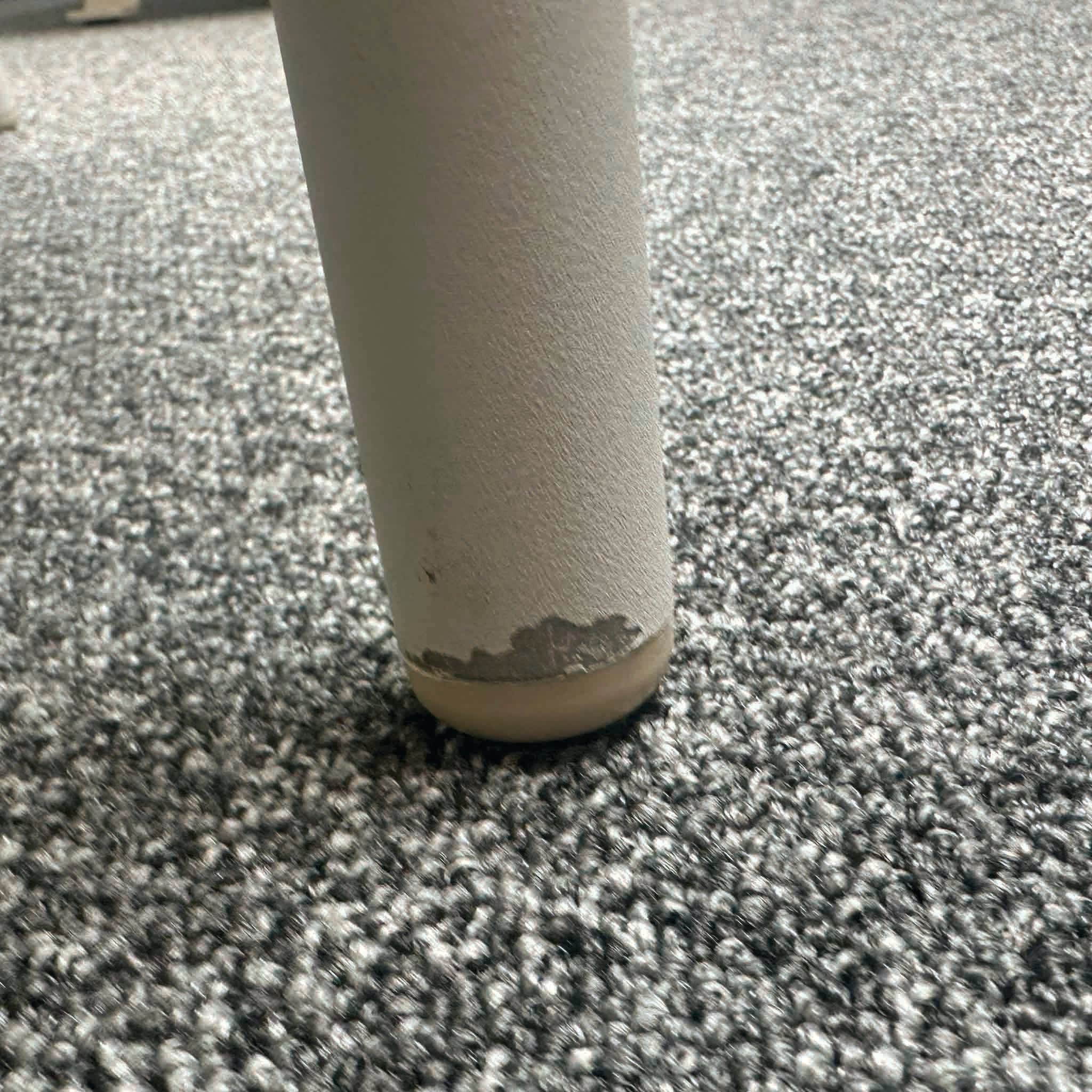 Close-up of a round table leg with chipped paint resting on a grey carpet; part of the distinctive Bernini 6 Seat Round Dining with Teak Table in Light Grey (Ex Display) range, now available at a special sale price.