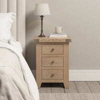 The Burford 3 Drawers Bedside Table in Natural Oak stands elegantly next to the bed, supporting a lamp, books, and a mug. Its pristine white bedding complements the table's rustic charm.