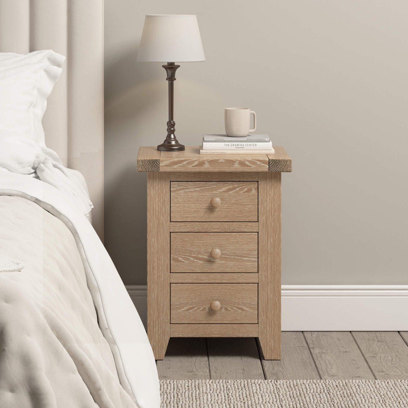 A natural oak Burford bedside table with three drawers holds a lamp and a mug, nestled next to a bed with crisp white bedding.