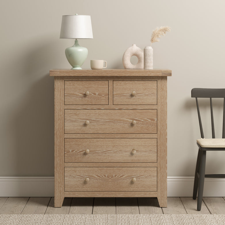 The Burford 2 Over 3 Chest of Drawers in Natural Oak showcases a lamp, mug, and vases elegantly placed on top, with a chair positioned beside it on the light carpeted floor, offering perfect storage solutions for any room.