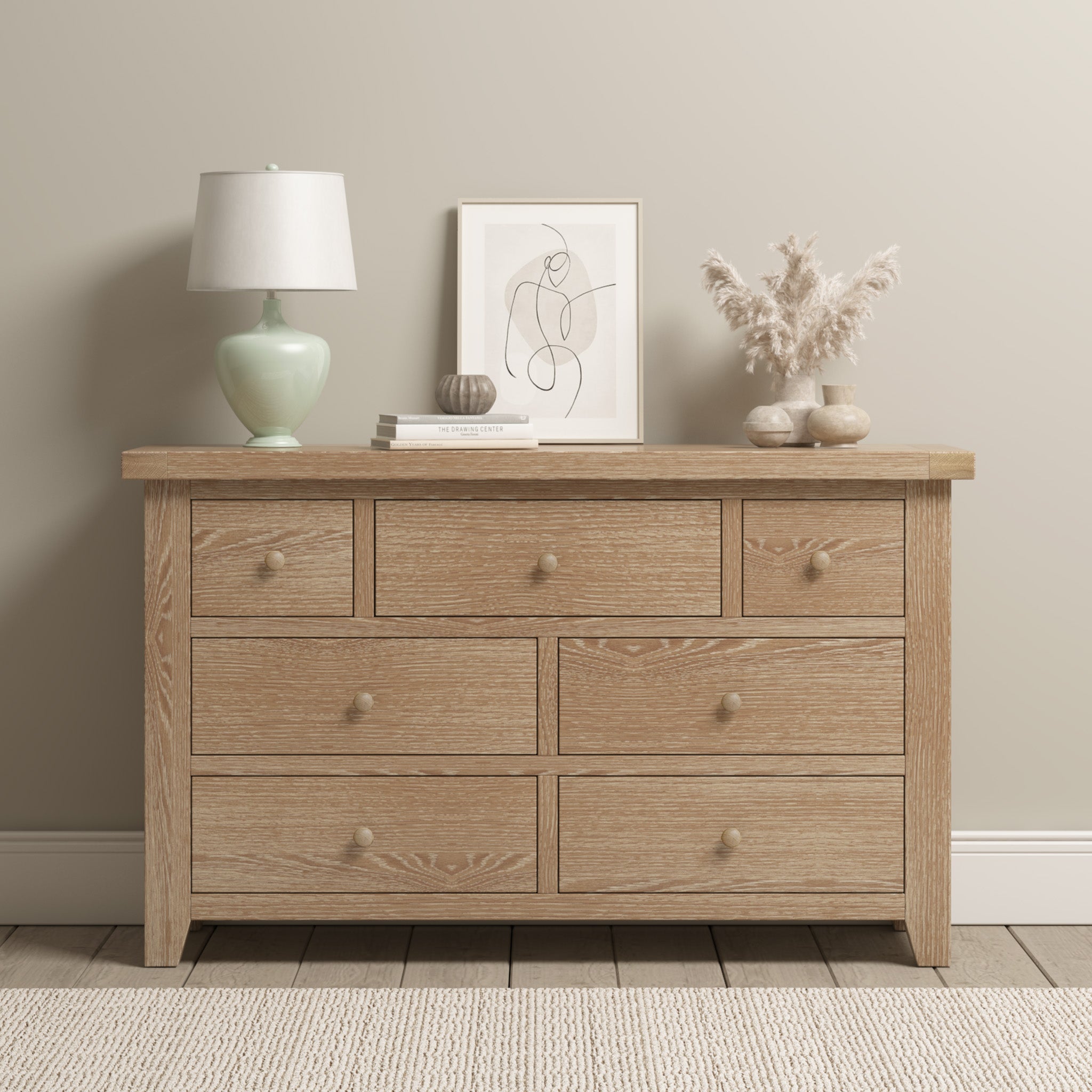 The Burford 3 Over 4 Chest of Drawers in Natural Oak provides generous storage and pairs well with the wooden dresser embellished with a lamp, books, framed art, and a vase filled with dried plants against a light wall.