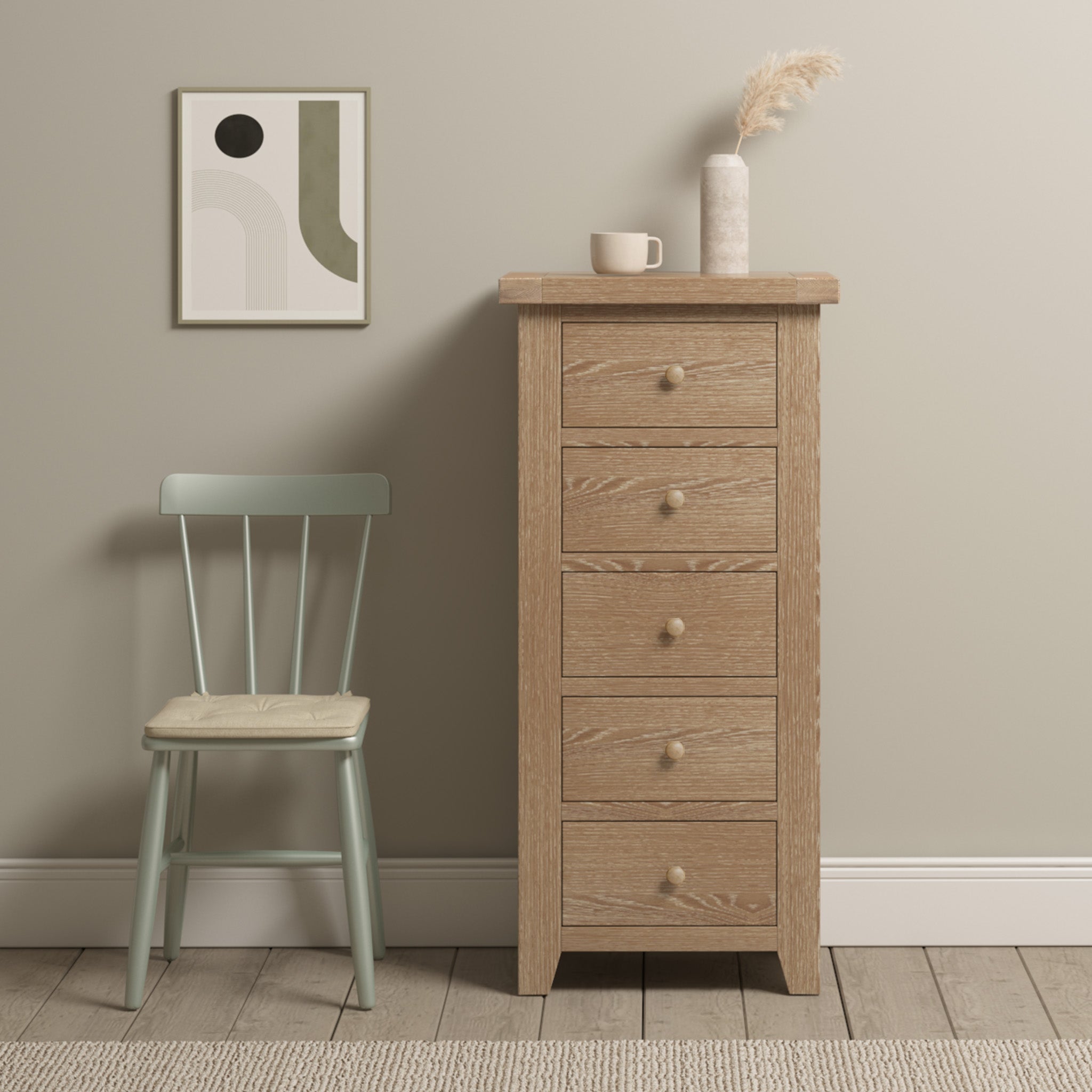 The Burford 5 Chest of Drawers in Natural Oak, decorated with a vase and mug, is placed beside a green chair and abstract wall art in a minimal room.