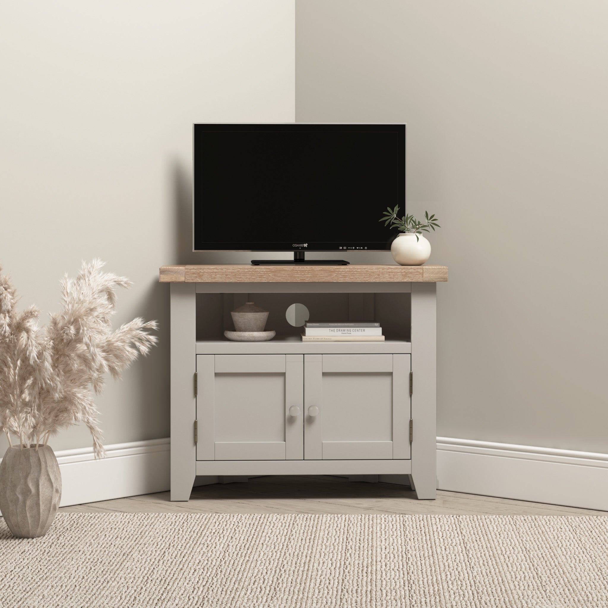 Pebble grey enhances the rustic charm of the Burford Corner Media Unit, where a sleek flat-screen TV sits alongside a small plant and books. On the left, pampas grass gracefully adorns a vase, completing this inviting setup.