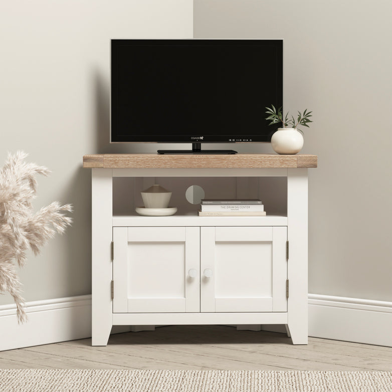 The Burford Corner Media Unit in Warm White showcases a small flat-screen TV, decorative plant, books, and pottery on its sleek white and wood cabinet, complete with efficient cable management.