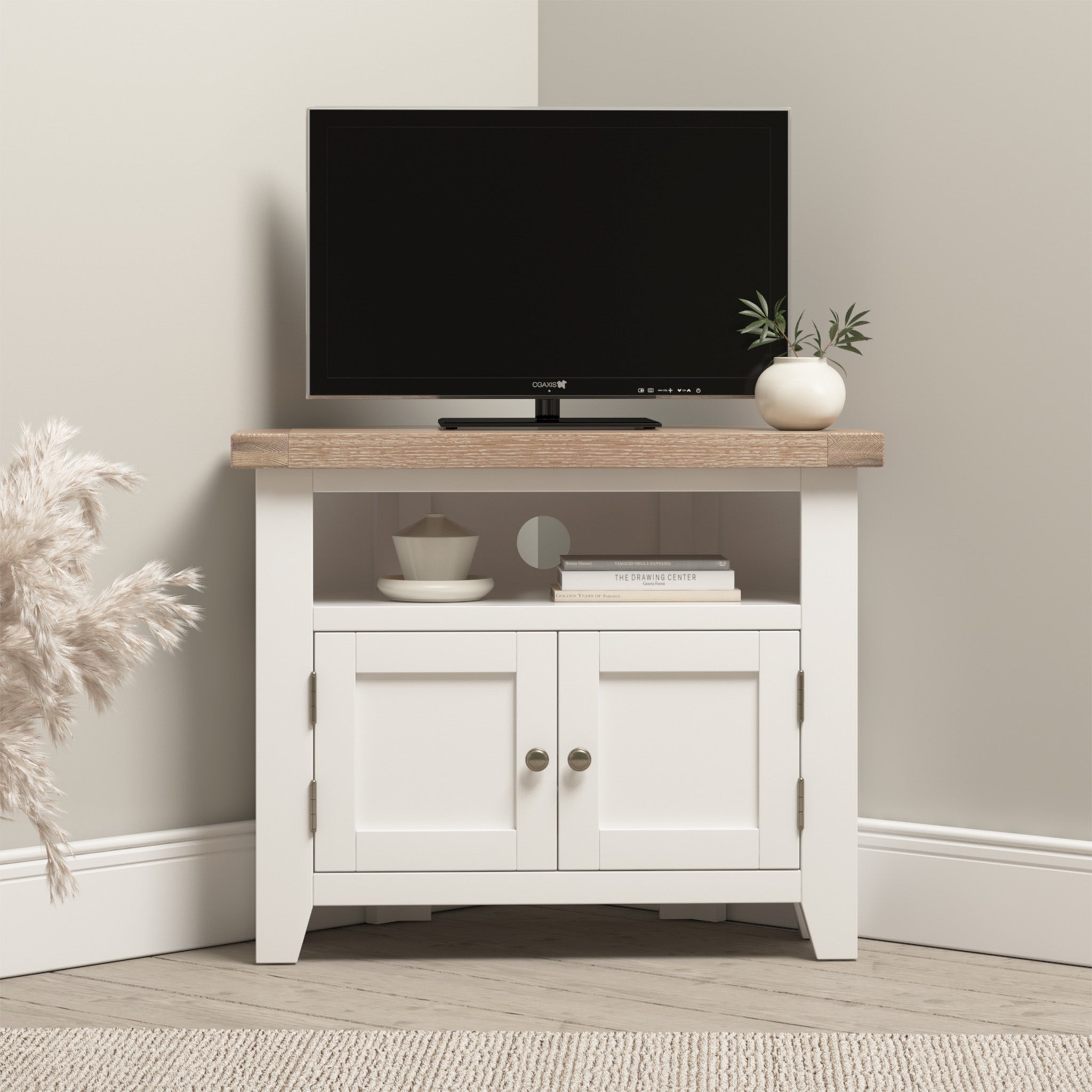 The Burford Corner Media Unit in Warm White elegantly showcases a small TV, decorative plant, books, and a bowl all neatly organized on its open shelf against the light-colored wall. The clever cable management ensures everything remains tidy and clutter-free.