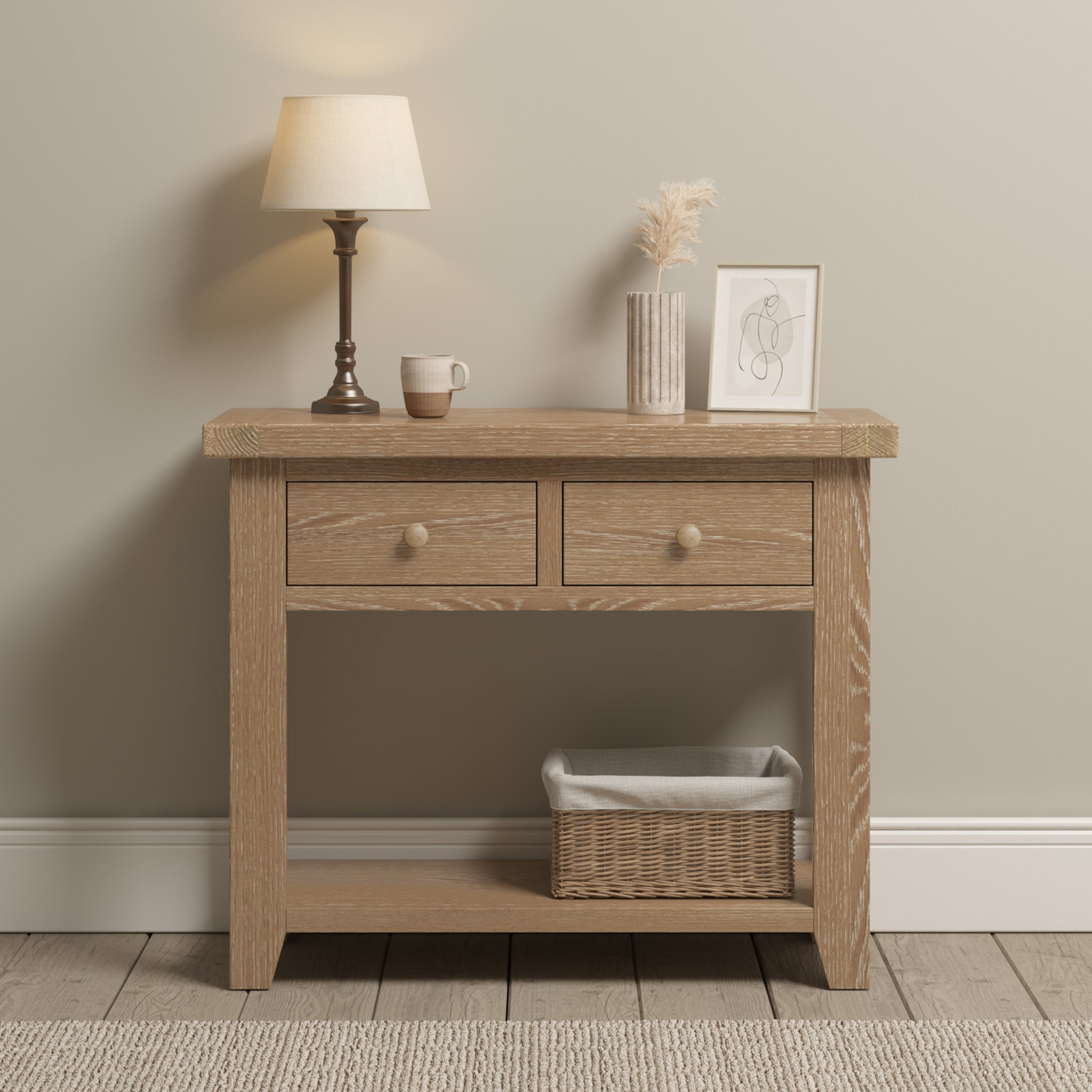 Experience the rustic charm of the Burford 2 Drawers Console Table in Natural Oak, adorned with a lamp, mug, vase, and photo on its surface. Below are two drawers and a wicker basket that sit elegantly atop a cozy rug.