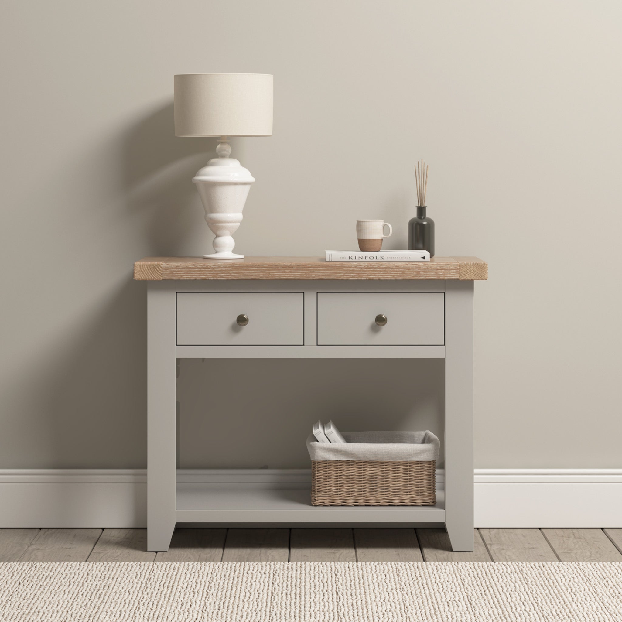 The Burford 2 Drawers Console Table in elegant Pebble Grey showcases a lamp, book, cup, and diffuser on its surface. A basket sits on the lower shelf against a beige wall, enhancing its charm.