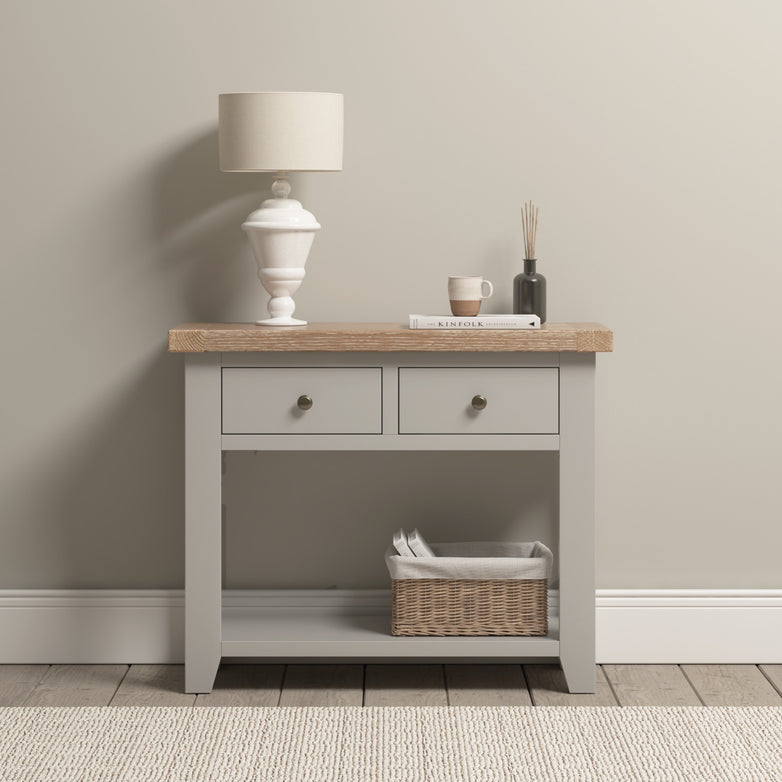 The Burford 2 Drawers Console Table in elegant Pebble Grey showcases a lamp, book, cup, and diffuser on its surface. A basket sits on the lower shelf against a beige wall, enhancing its charm.