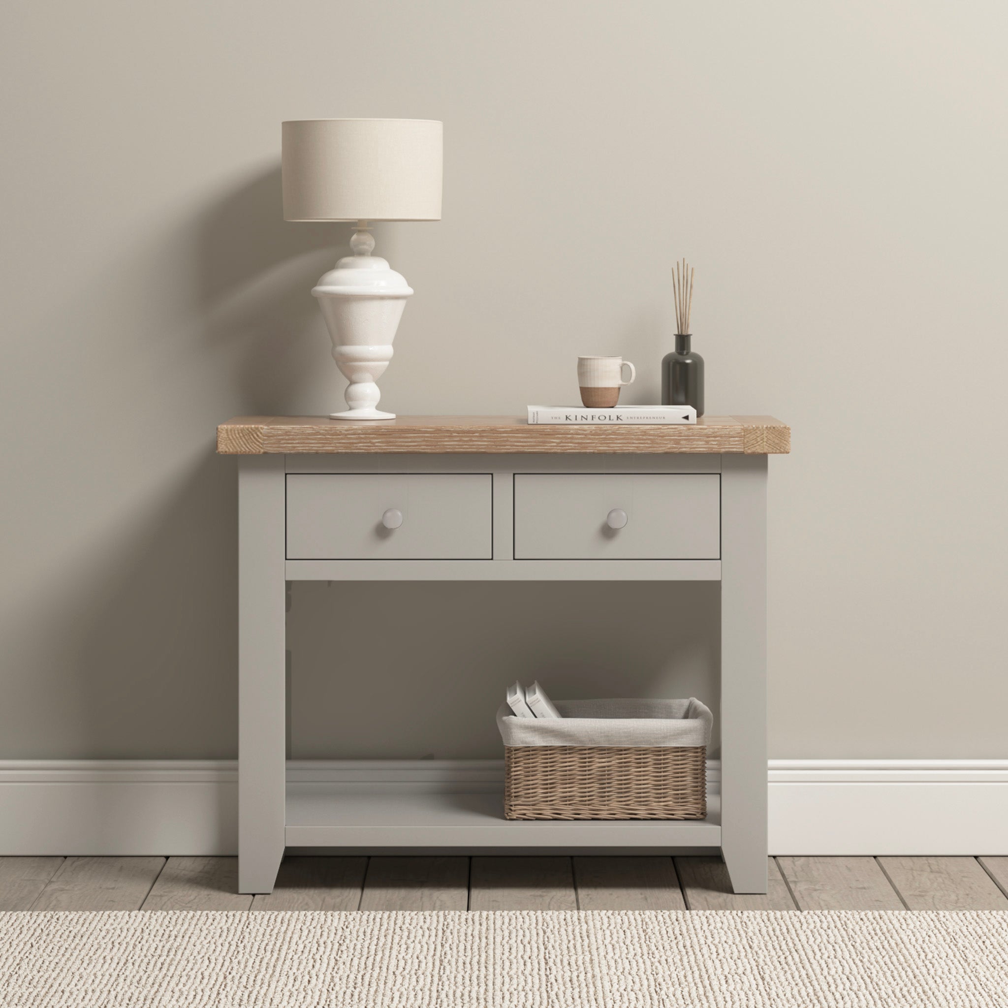 A Burford 2 Drawers Console Table in pebble grey stands against a neutral wall, adorned with a lamp, books, and a candle. A basket nestles underneath, adding charm to its elegant design.