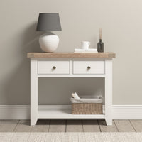 A Burford 2 Drawers Console Table in warm white displays a vase, lamp, mug, and basket against a light-colored wall. Its natural oak accents harmonize beautifully with the serene backdrop.