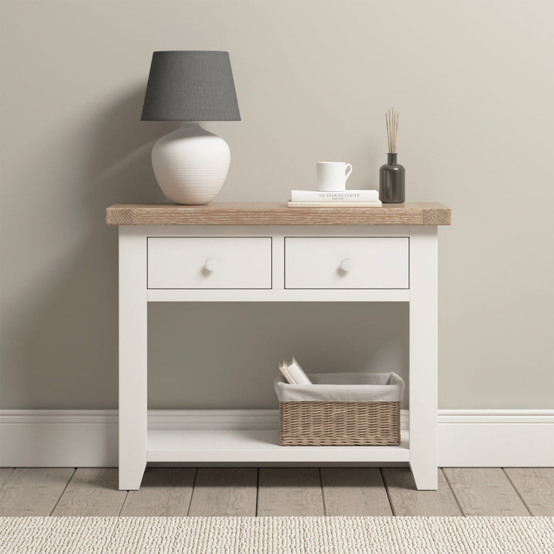 The Burford 2 Drawers Console Table in warm white is beautifully accented by a lamp, books, a mug, and a basket underneath. Its natural oak details enhance the elegance of any wooden floor setting.