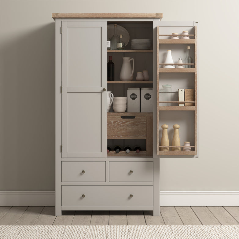 Open pantry cabinet with jars, dishes, and kitchen items on shelves, drawers below, showcasing storage and organization in a light-colored room. The Burford Double Larder Unit in Pebble Grey adds elegance and warmth to your kitchen space.