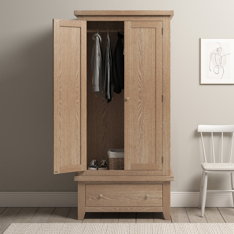 The Burford Double Wardrobe with Drawer in Natural Oak stands with its doors open, showcasing clothes and a basket inside. Its natural oak finish enhances its elegance. Nearby, a chair and artwork complement this solid oak wardrobe, which perfectly blends style and functionality.