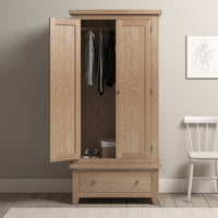 The Burford Double Wardrobe with Drawer in Natural Oak, shown with open doors displaying jackets inside and a convenient basket below, stands next to a white chair and abstract wall art. Its natural oak finish infuses warmth, effortlessly complementing any decor style.