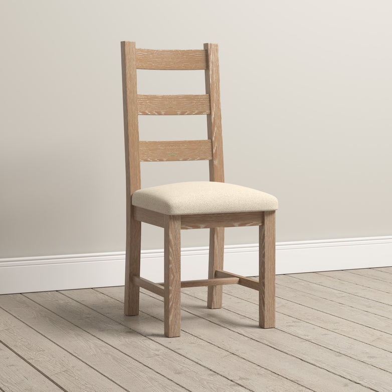 The Burford Set of 2 Ladderback Dining Chairs with Fabric Seat in Natural Oak showcases a natural oak finish and an upholstered fabric seat that harmonizes beautifully with its wooden frame. Placed on a light wood floor against an off-white wall, these chairs seamlessly blend elegance with comfort.