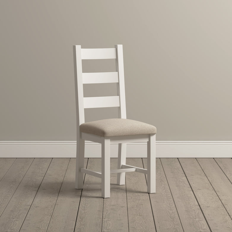 A pair of Burford Ladderback Dining Chairs in warm white, featuring fabric seats in beige, elegantly rests on a wooden floor against a neutral wall. Their refined design amplifies the natural oak finish, providing timeless charm to any dining area.